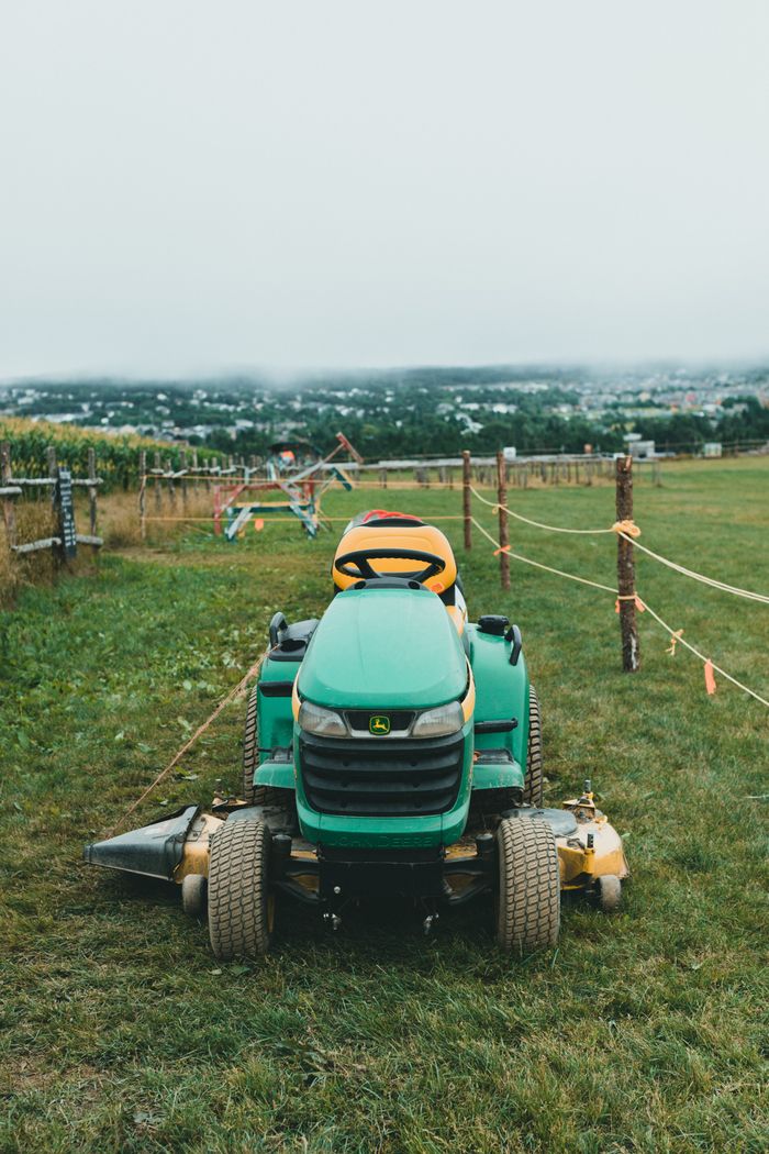 A green riding lawn mower sits on a vast grassy field with rustic fencing in a tranquil rural setting.