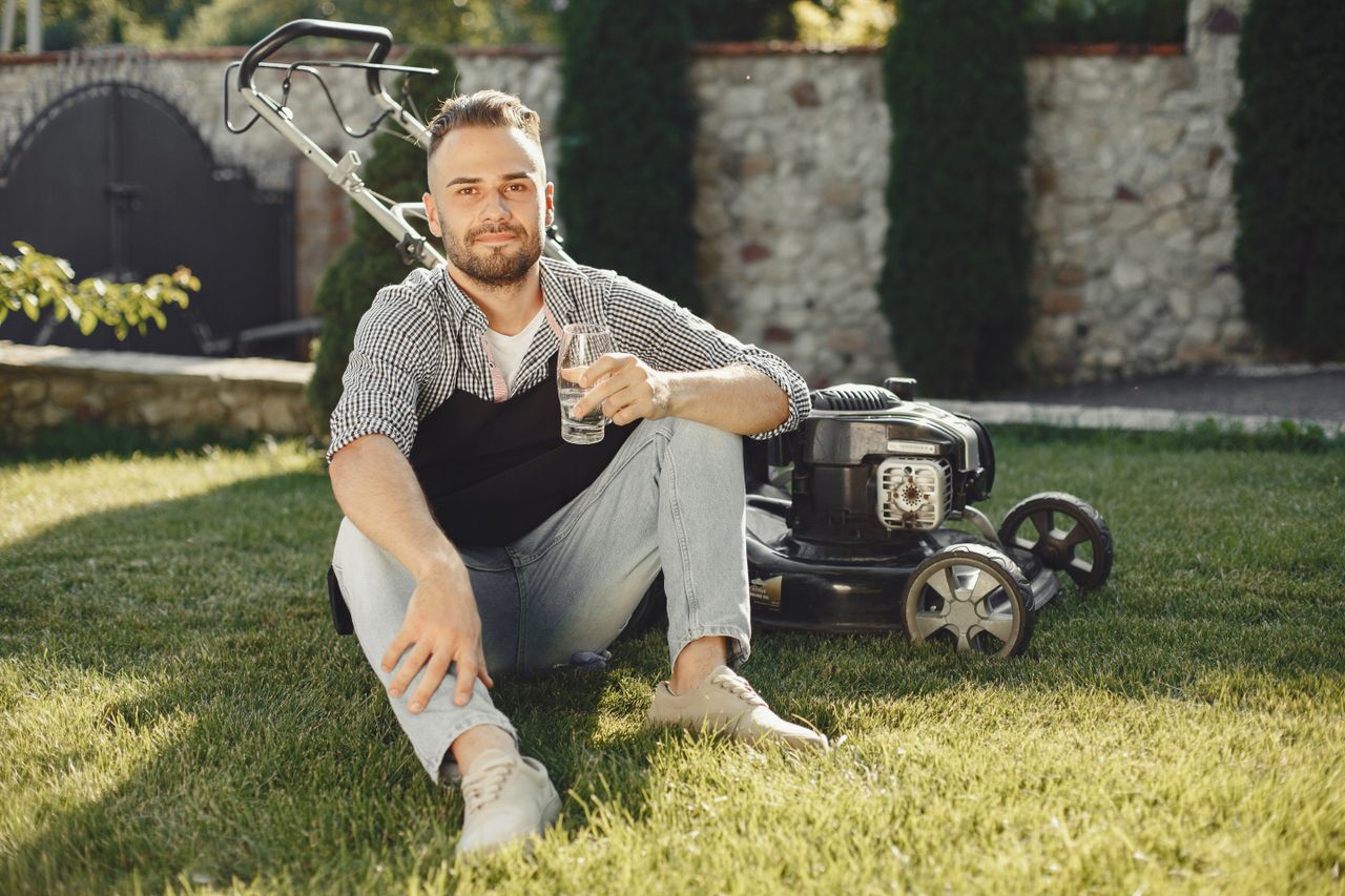 Man sitting on grass with drink, beside lawn mower on a sunny day.